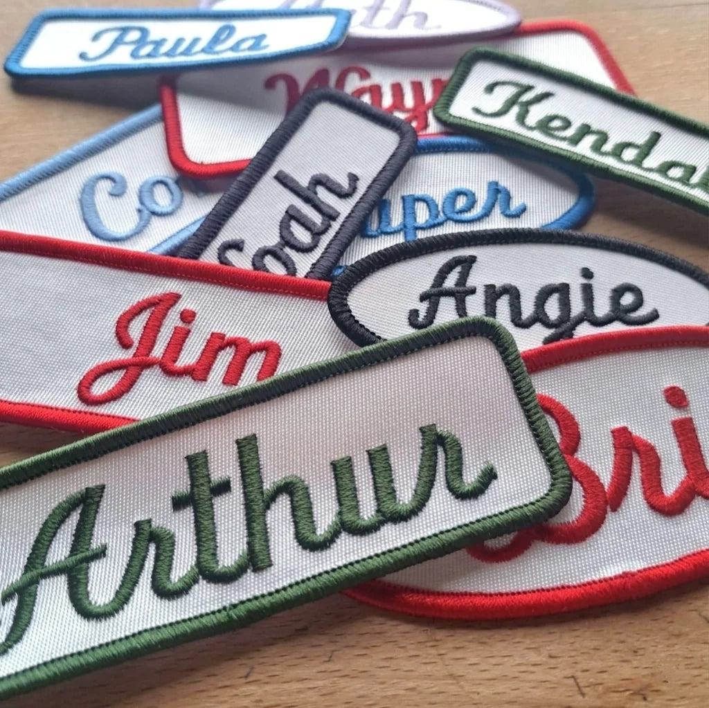 A pile of embroidered name patches on a wooden surface. The patches are white with colorful embroidered fonts and borders 